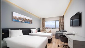 Une chambre d’hôtel avec deux lits, un bureau, une chaise et une grande fenêtre donnant sur le paysage urbain.