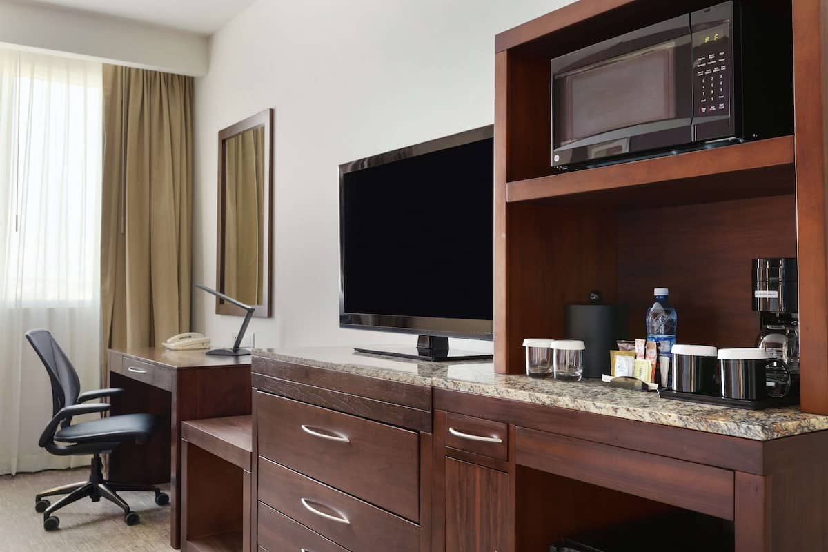A hotel room with a wooden desk, a flat-screen TV, a microwave, a coffee maker, and a water bottle.