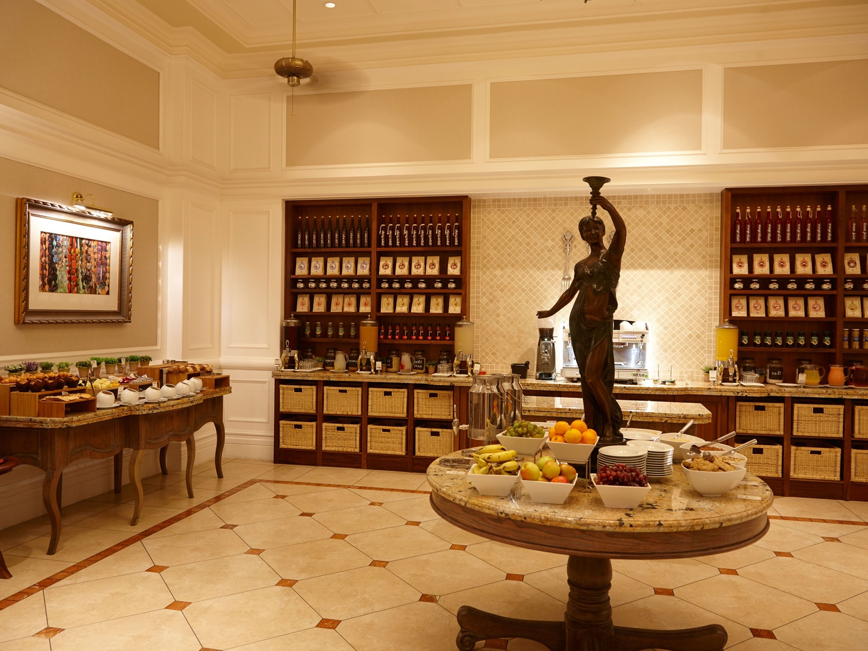 A spacious room with a central round table displaying fruits and a statue, surrounded by shelves with bottles and jars.