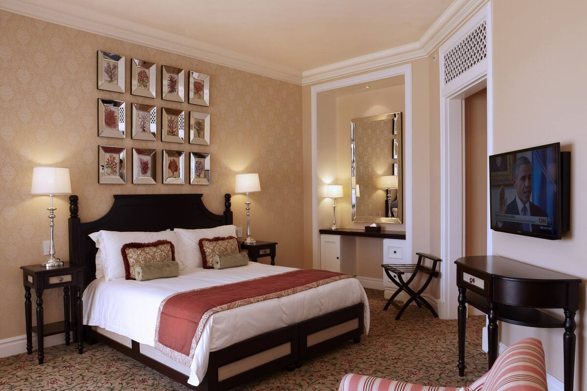 Un cuarto de hotel con una cama grande, dos mesitas de noche con lámparas, un escritorio con silla, un espejo amplio y cuadros enmarcados en la pared.