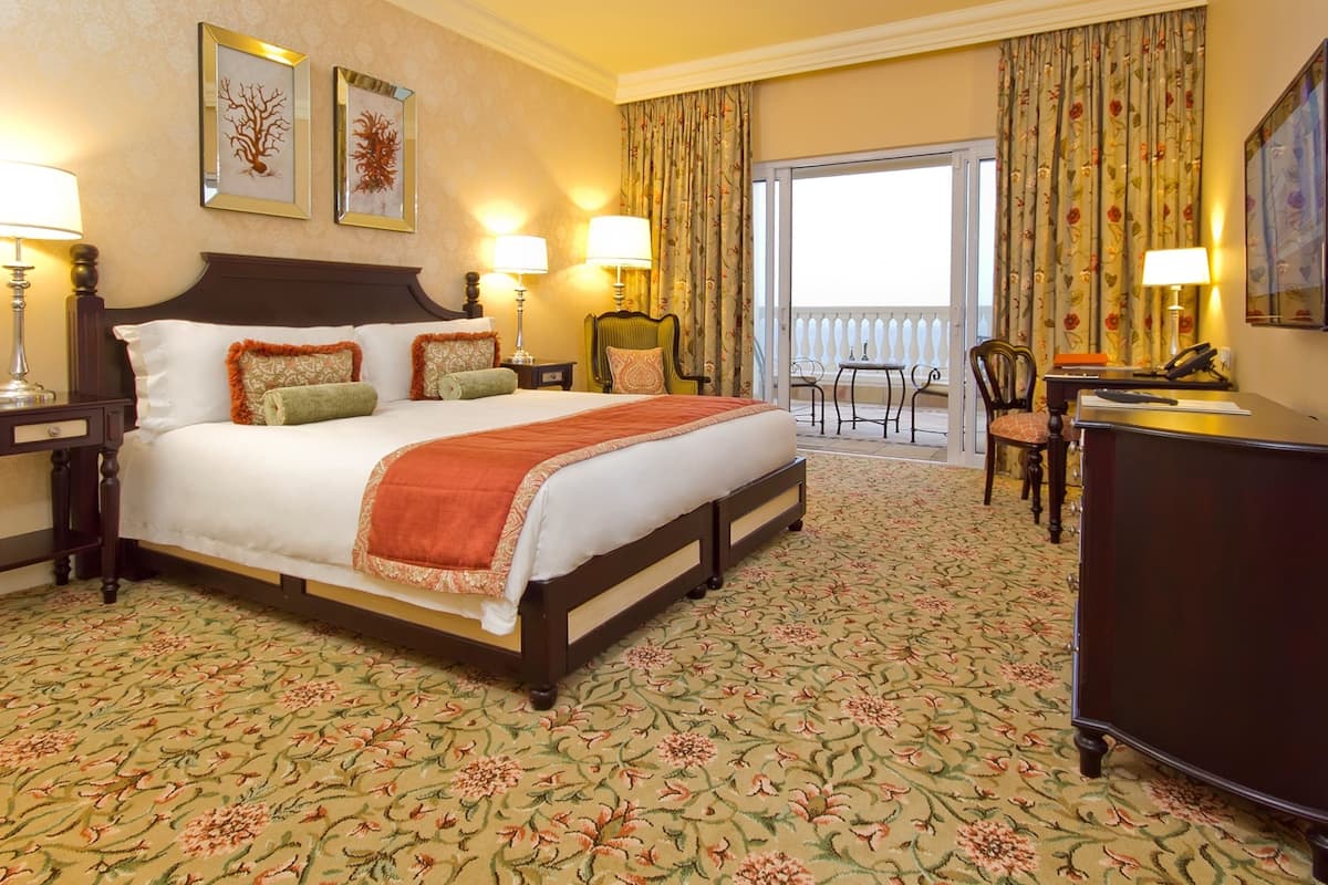 Habitación de hotel con una cama grande, dos mesitas de noche con lámparas, un escritorio con una silla, un balcón con cortinas y un ventanal amplio.