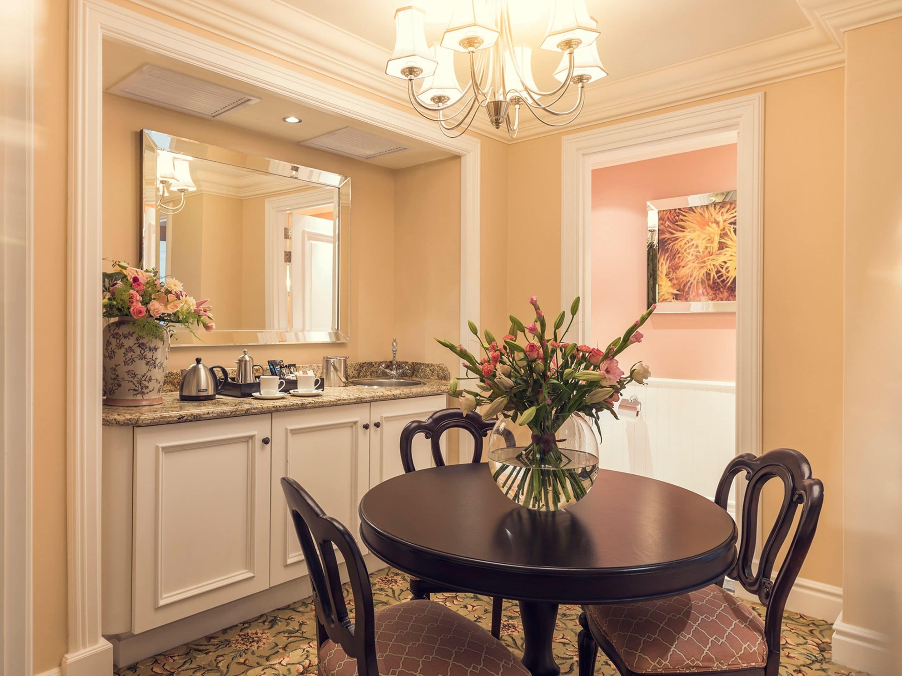 A dining area with a round table, chairs, a vase with flowers, a kitchen counter with a kettle, and a large mirror above the counter.