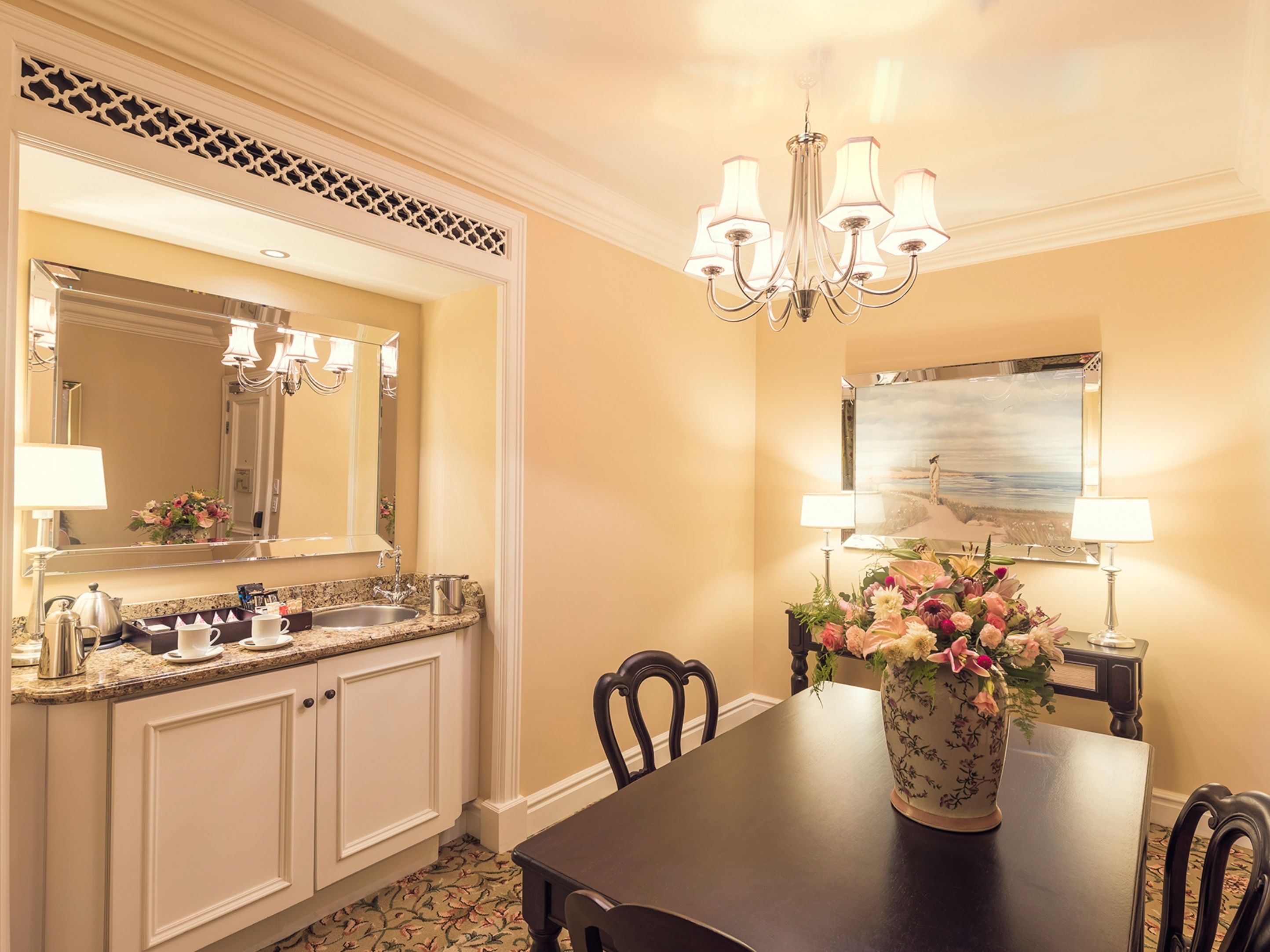 A dining area with a chandelier, a large mirror, and a vase of flowers on a table.