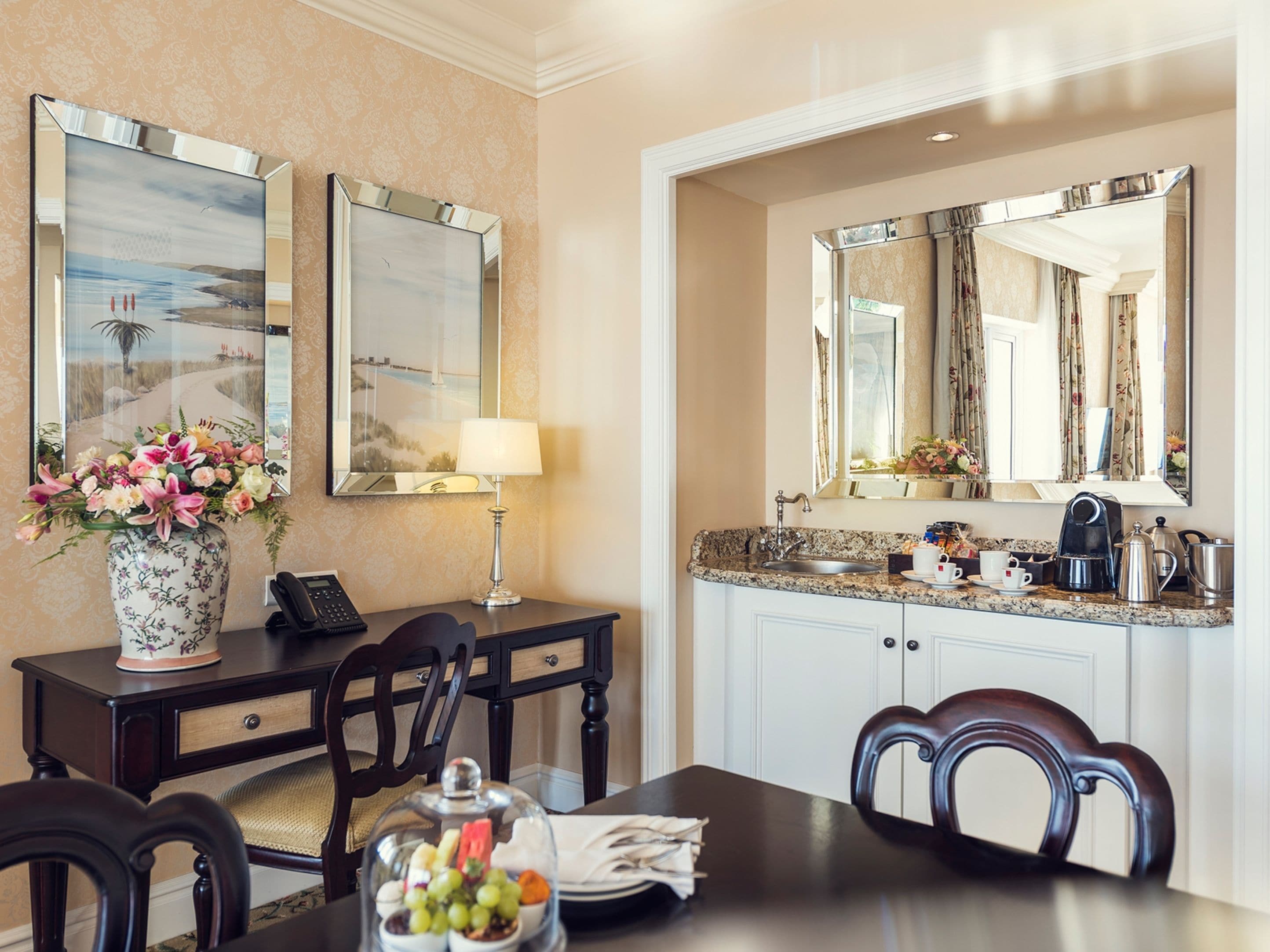 Habitación de hotel con zona de comedor, un escritorio con teléfono, lámpara, un jarrón con flores, frutero y un espejo grande.