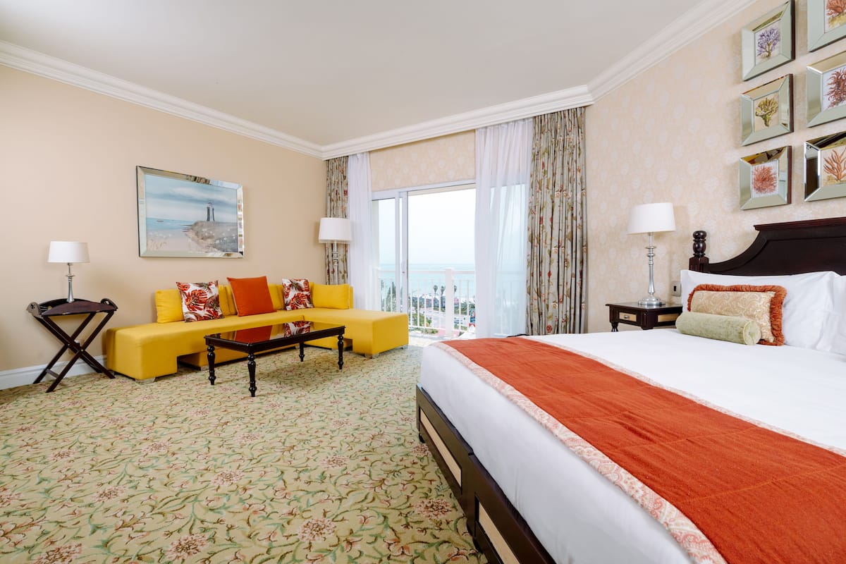Un cuarto de hotel con una cama grande, un sofá amarillo, una mesita de café y vista al mar a través de la ventana.