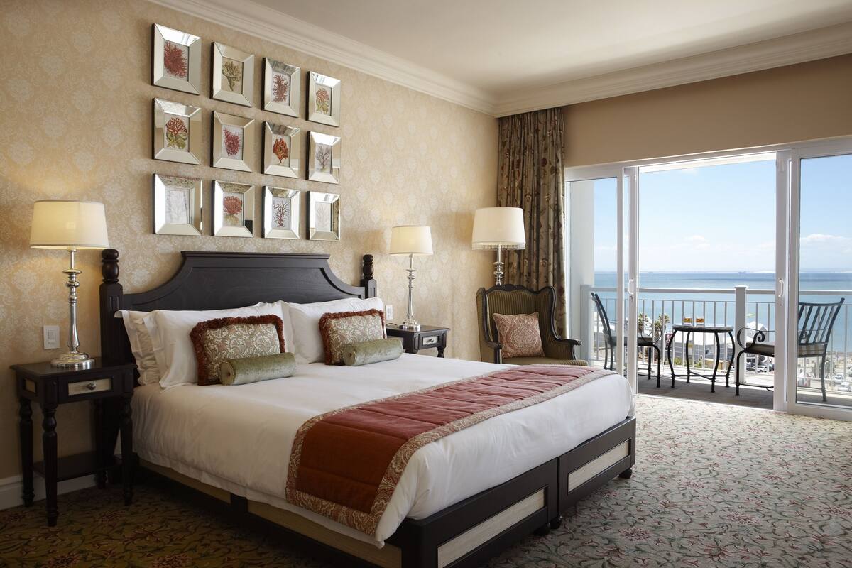 Habitación de hotel con una cama grande, dos mesitas de noche, una silla, un balcón con vistas al mar y cuadros en la pared.