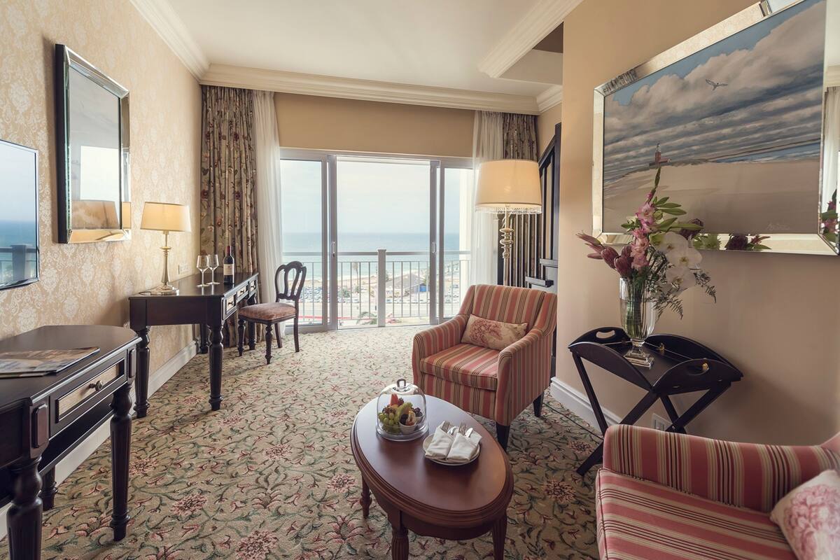 Habitación de hotel con balcón, una mesa de comedor con frutero, un sillón a rayas y un escritorio con lámpara.