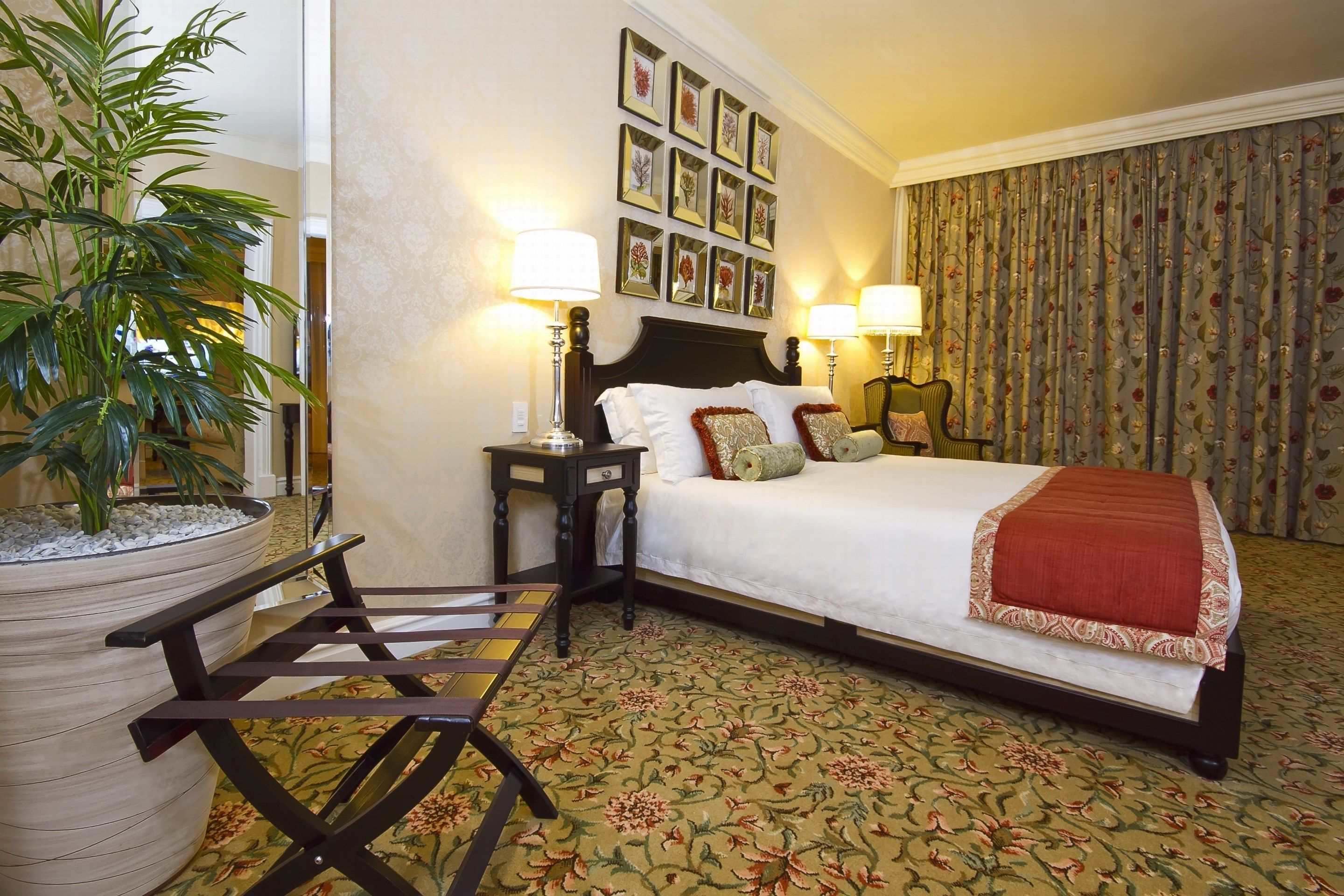 Hotelkamer met een bed, twee nachtkastjes met lampen, een grote potplant, een houten stoel en een bloemenkleed.