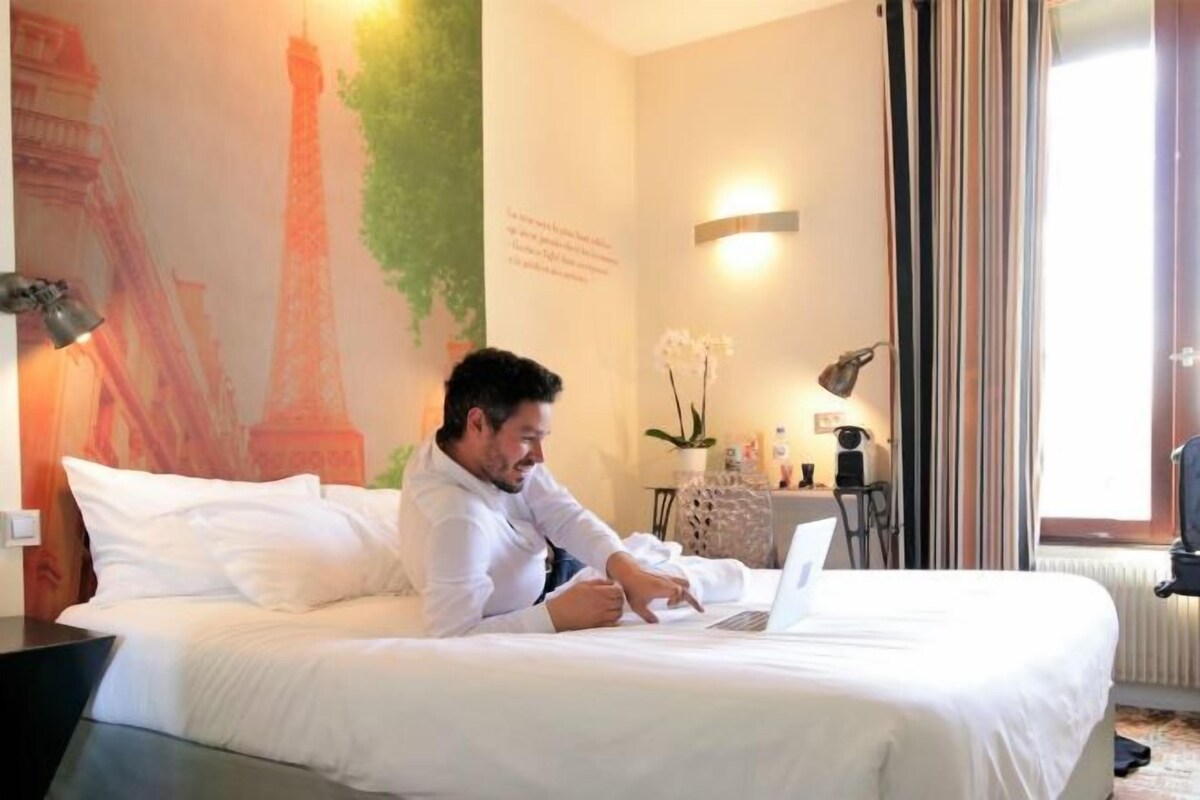 Habitación de hotel con cama, computadora portátil, pared con mural, lámpara y ventana con cortinas.