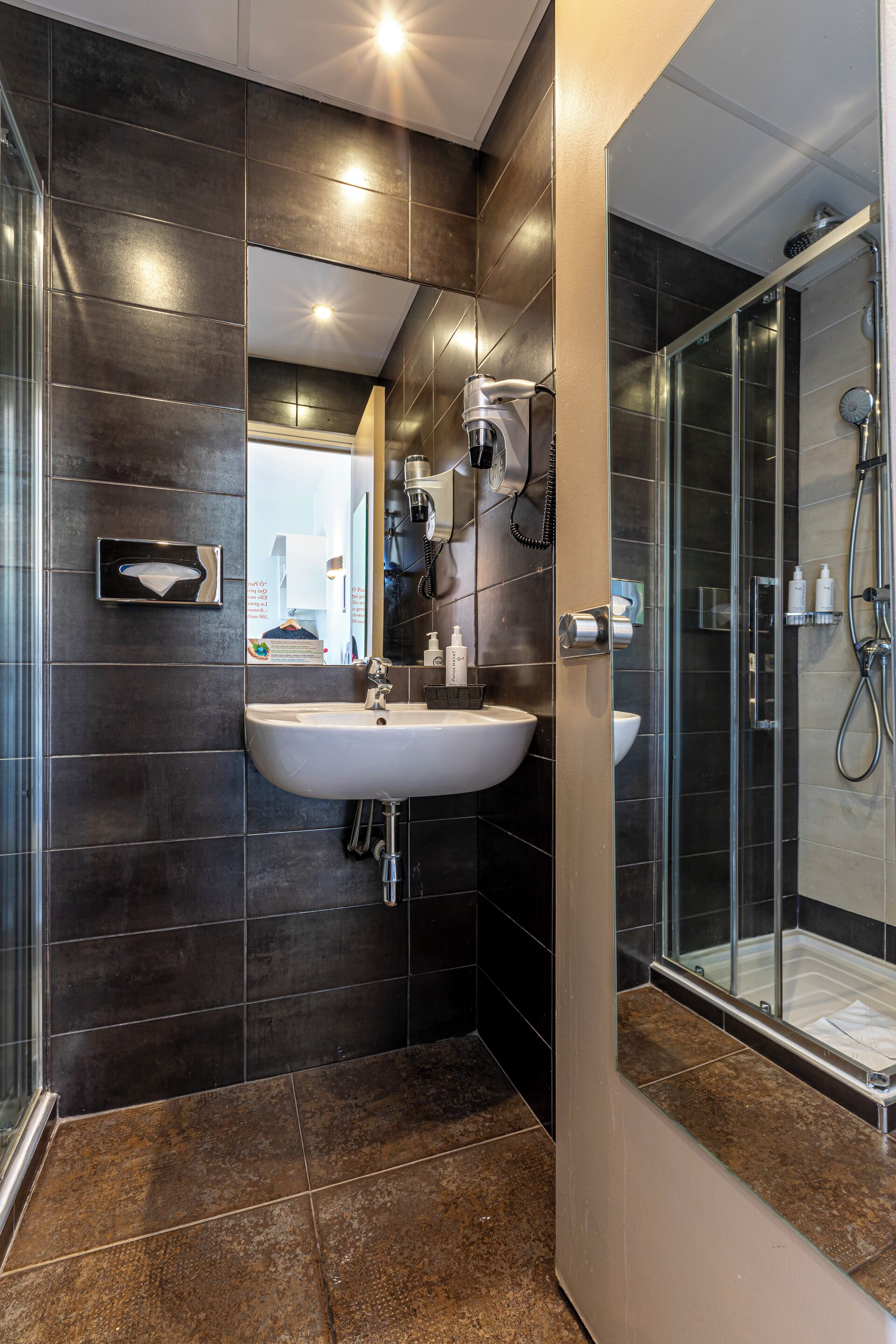 Un bagno moderno con piastrelle scure, un lavandino bianco e un box doccia in vetro.
