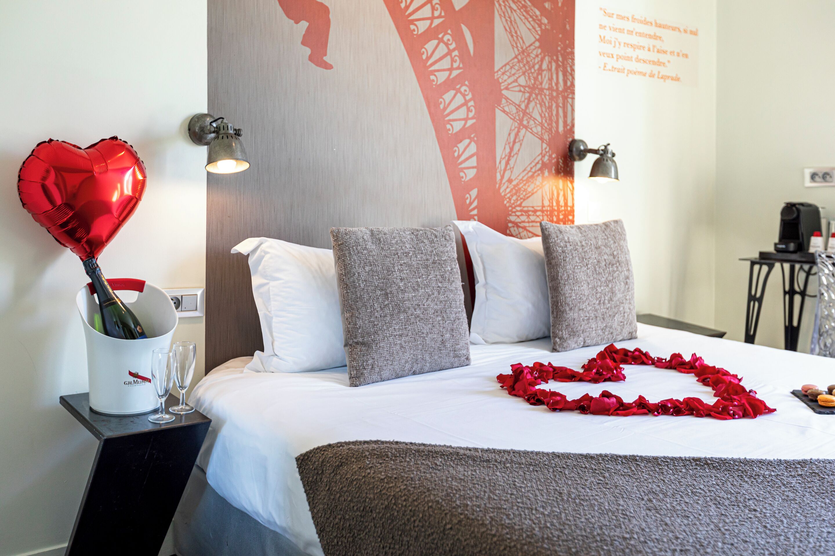 Ein Hotelzimmer mit einem Bett, Kissen, einem Nachttisch mit Champagnerflasche und Gläsern sowie einem dekorativen Herzballon.