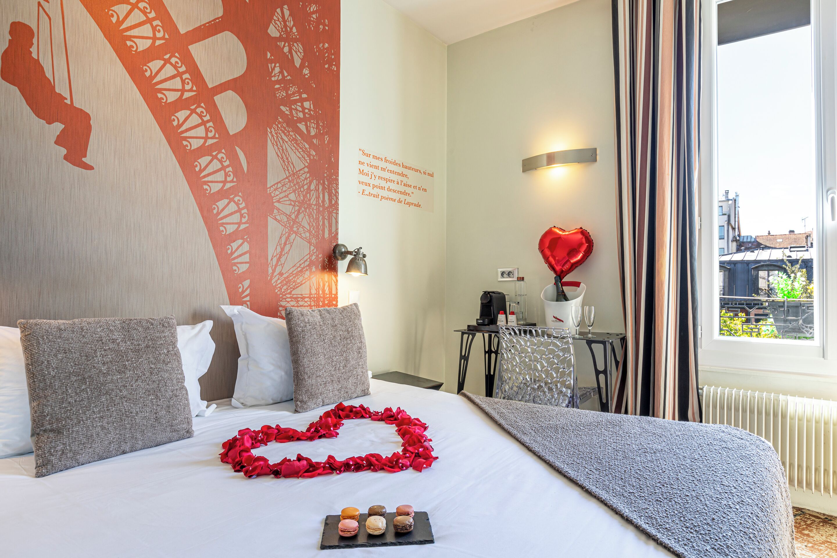 A hotel room with a bed, pillows, a heart-shaped decoration, a tray of macarons, a red balloon, and a view of the city through the window.