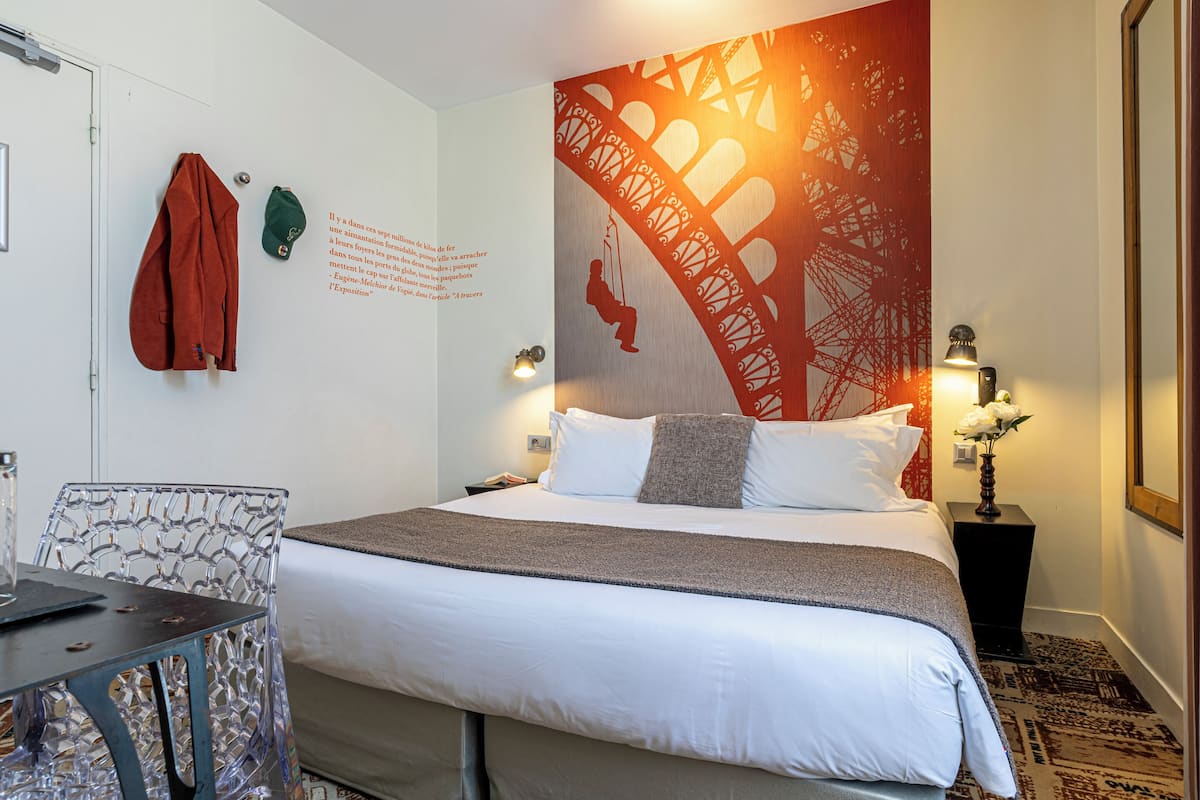 A hotel room with a neatly made bed, a chair, a table, and a wall with a decorative pattern.