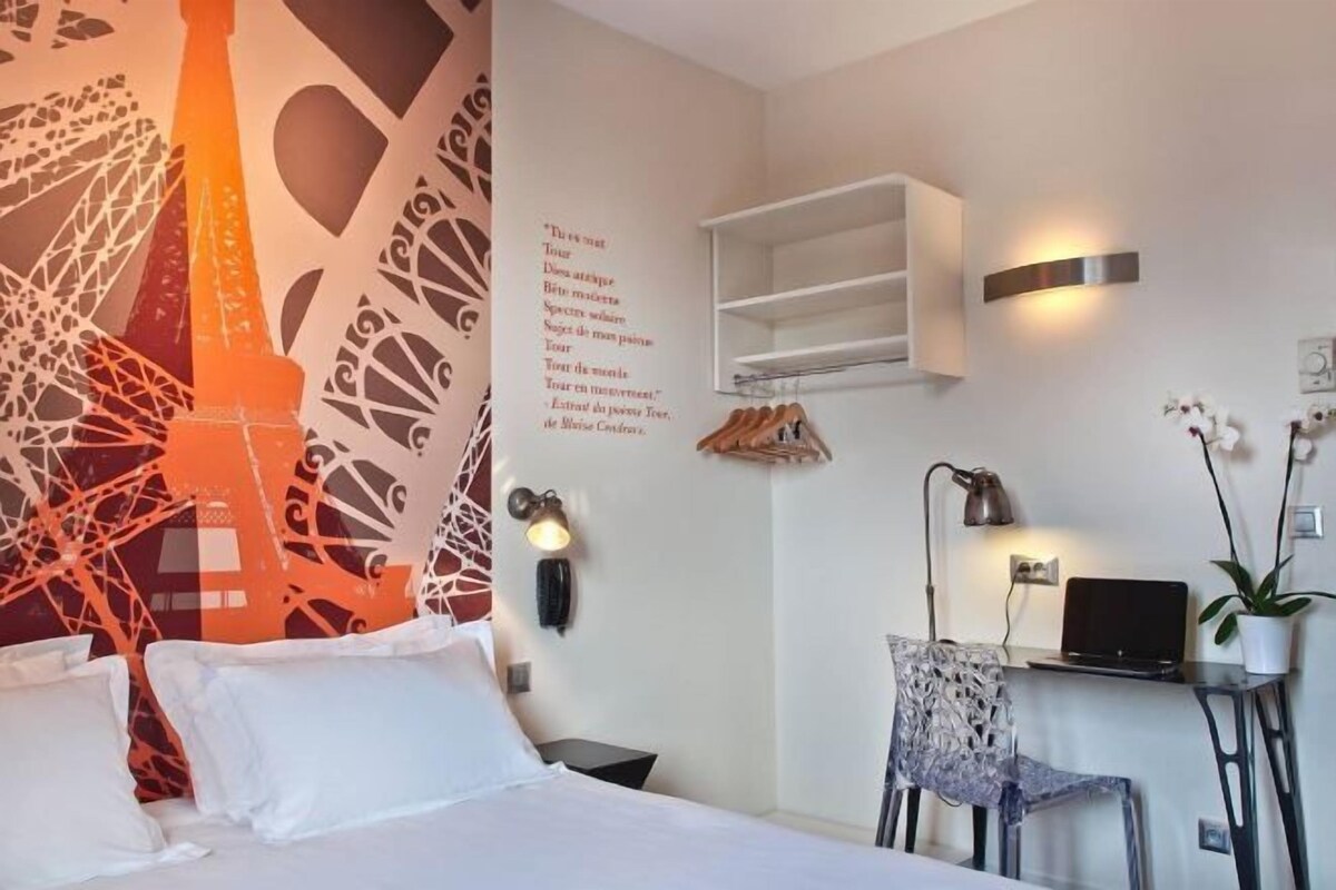Habitación de hotel con cama, escritorio con laptop, silla, estante montado en la pared y un adorno decorativo.