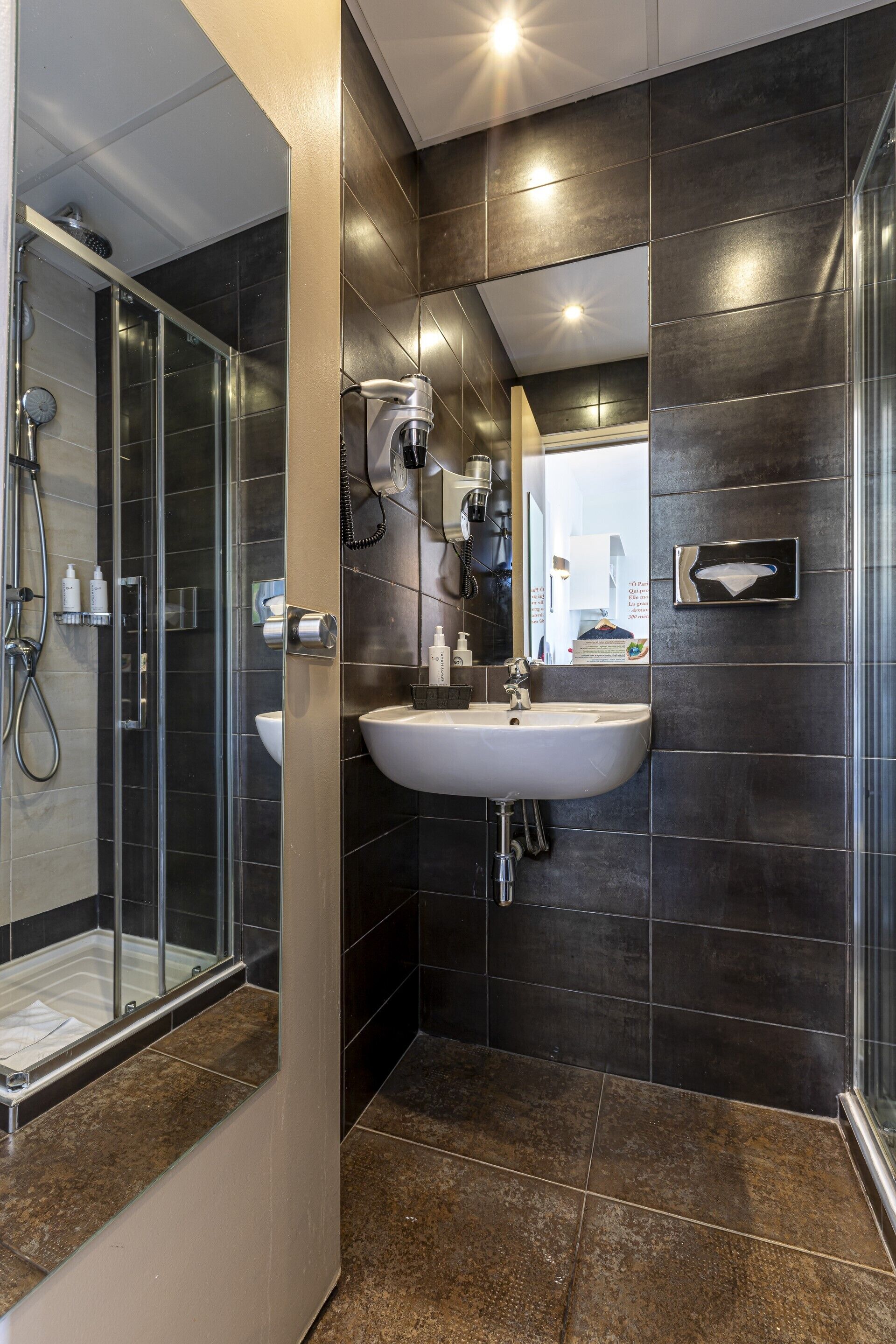 A modern bathroom with a glass shower enclosure, a white sink, and dark tiled walls.