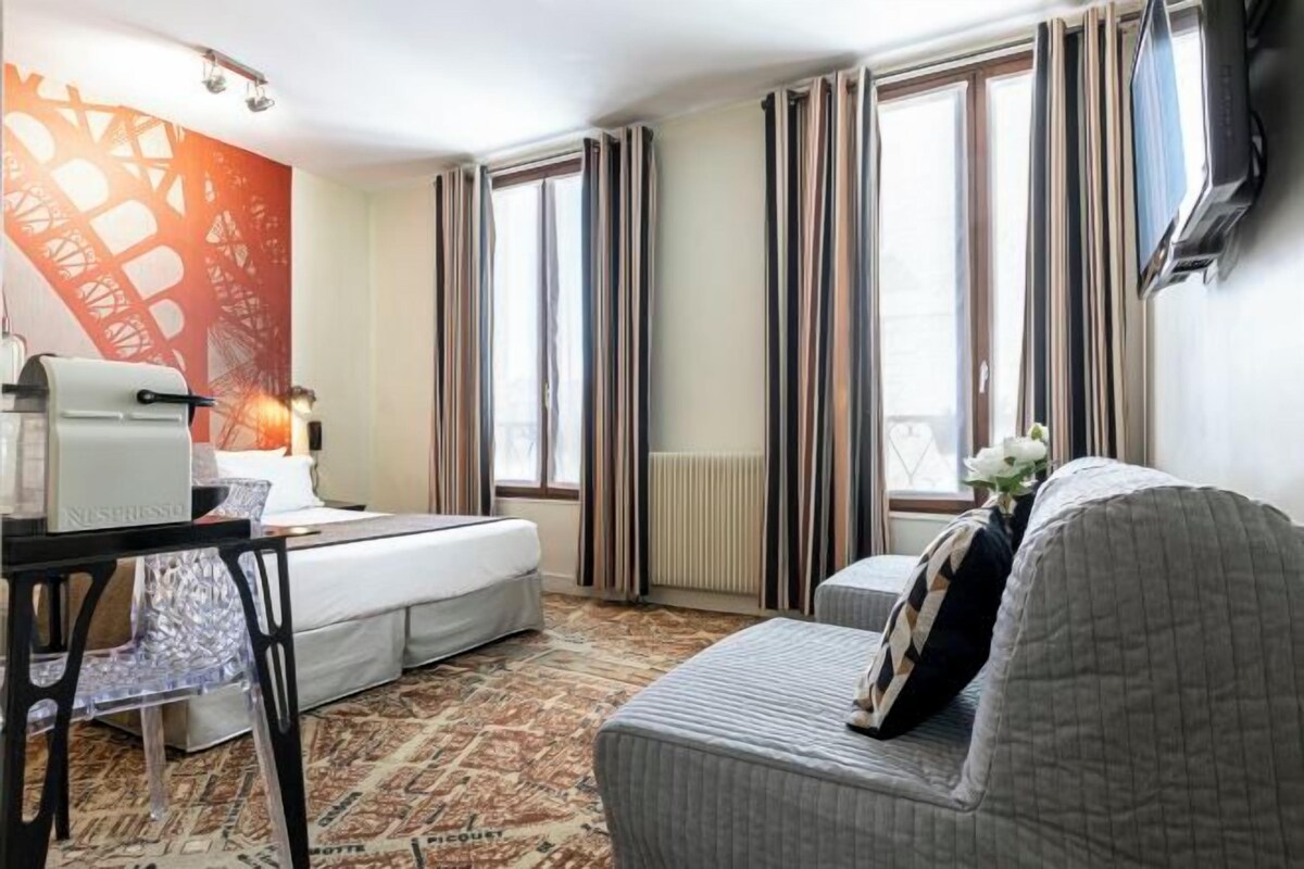 Habitación de hotel con cama, escritorio con cafetera, silla, sofá y ventana grande con cortinas.