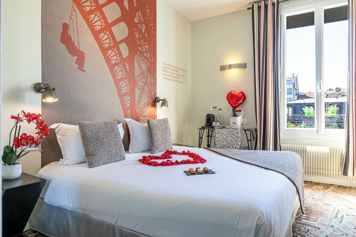 A hotel room with a large bed, a wall mural, a small table with a lamp, and a window with curtains.