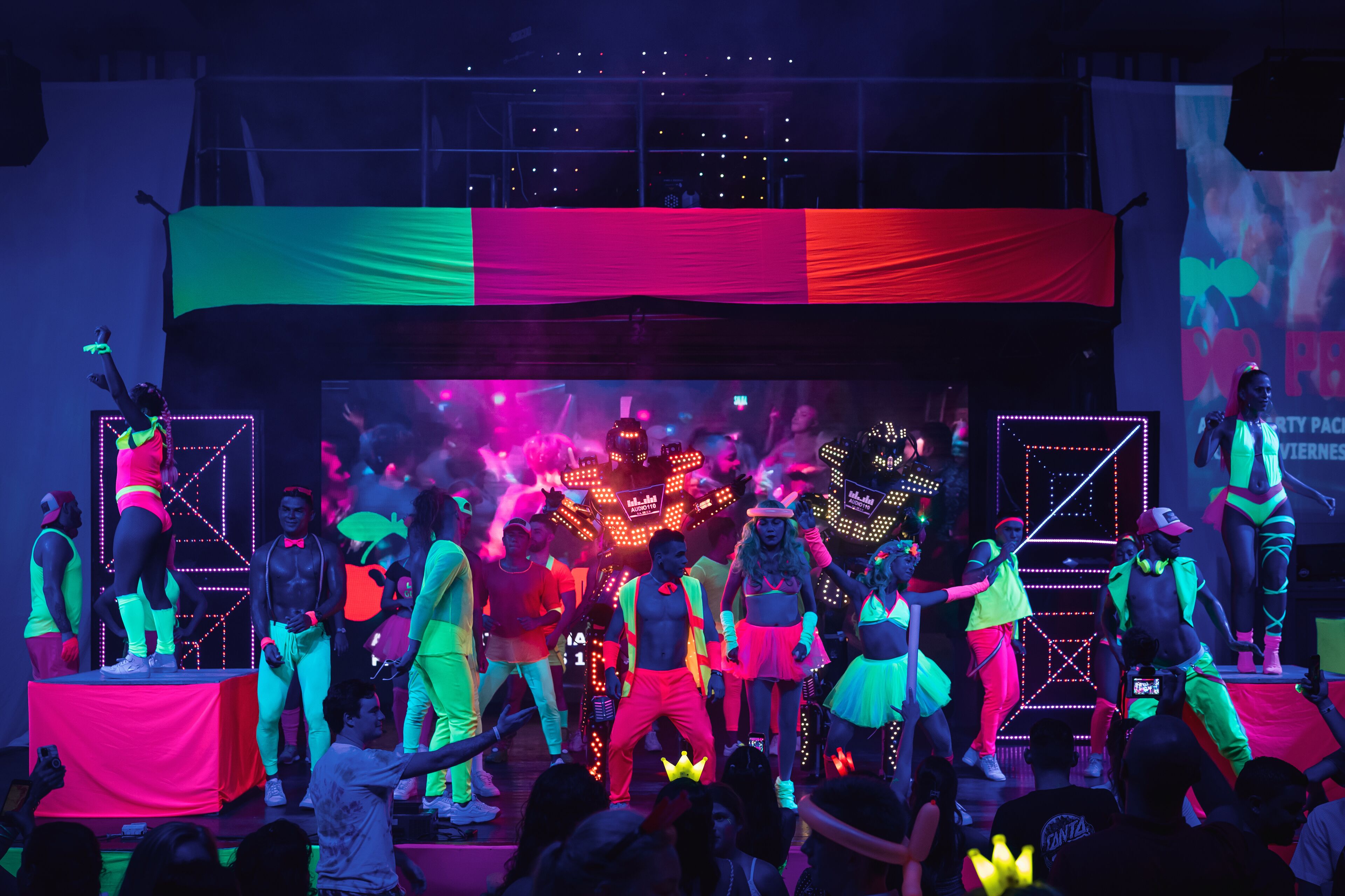 A stage with performers in neon outfits, a large LED screen, and a crowd of spectators.