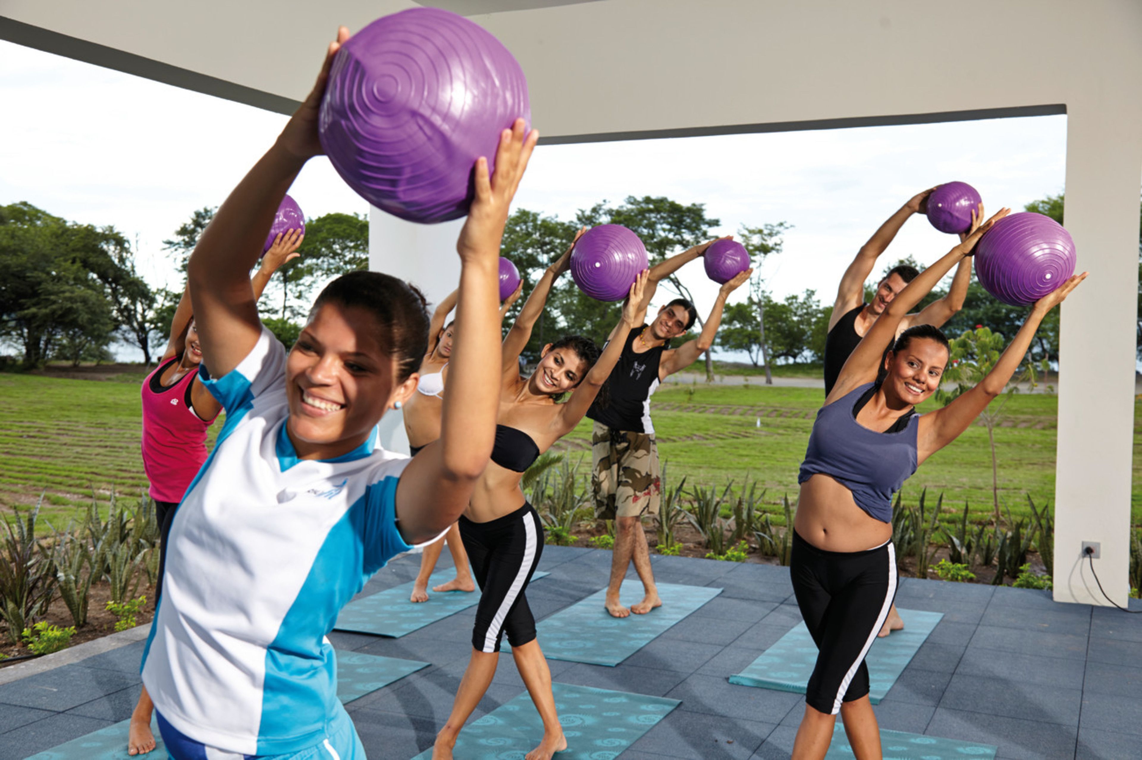 Eine Gruppe Menschen trainiert mit violetten Gymnastikbällen auf Yogamatten im Freien, in einem weitläufigen, offenen Bereich mit Blick auf eine Wiese.