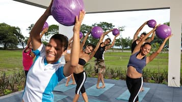 A group of people exercising with purple exercise balls on outdoor yoga mats in a spacious, open-air area with a view of a grassy field.