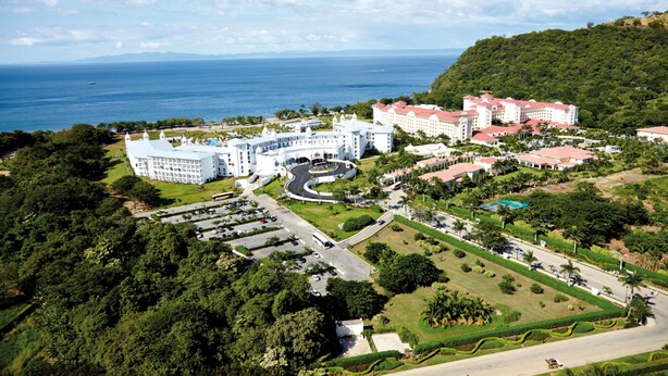 An aerial view of a resort with multiple buildings, a central roundabout, and a coastline in the background. - Hotel Riu Palace Costa Rica - All Inclusive (Sardinal)