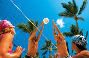 A beach volleyball game with players reaching for the ball, palm trees, and a clear blue sky.