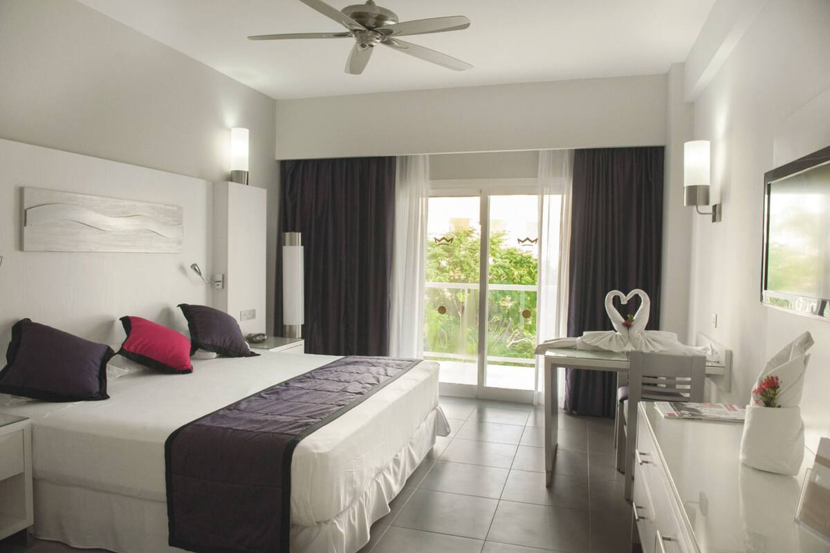Habitación de hotel con cama, escritorio, silla, ventilador de techo y balcón con cortinas.