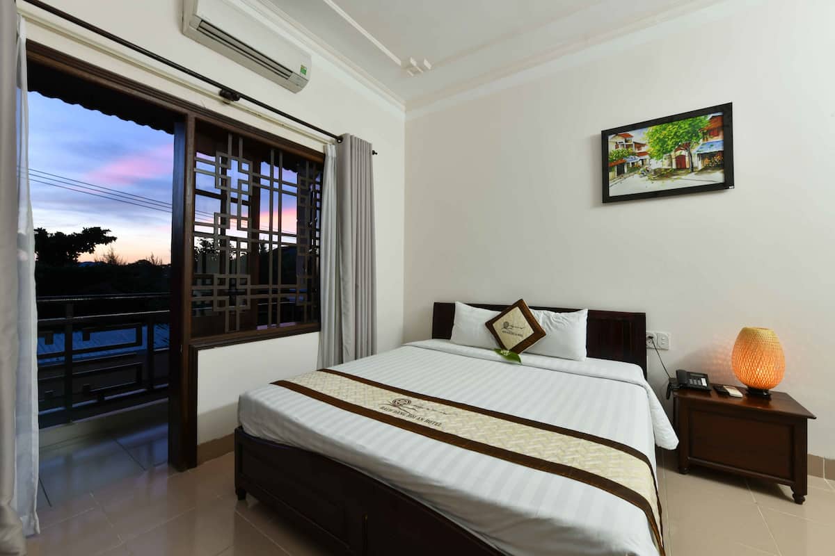 A hotel room with a bed, a nightstand with a lamp, a painting on the wall, and a view of the outside through a window.