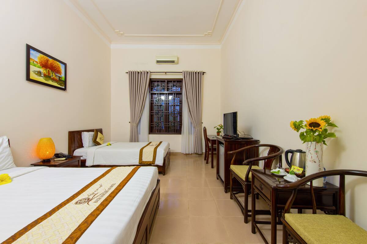 A hotel room with two beds, a desk, a chair, a television, and a vase with sunflowers.