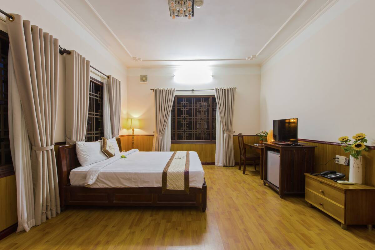 A hotel room with a wooden bed, a desk with a TV, a lamp, and a window with curtains.