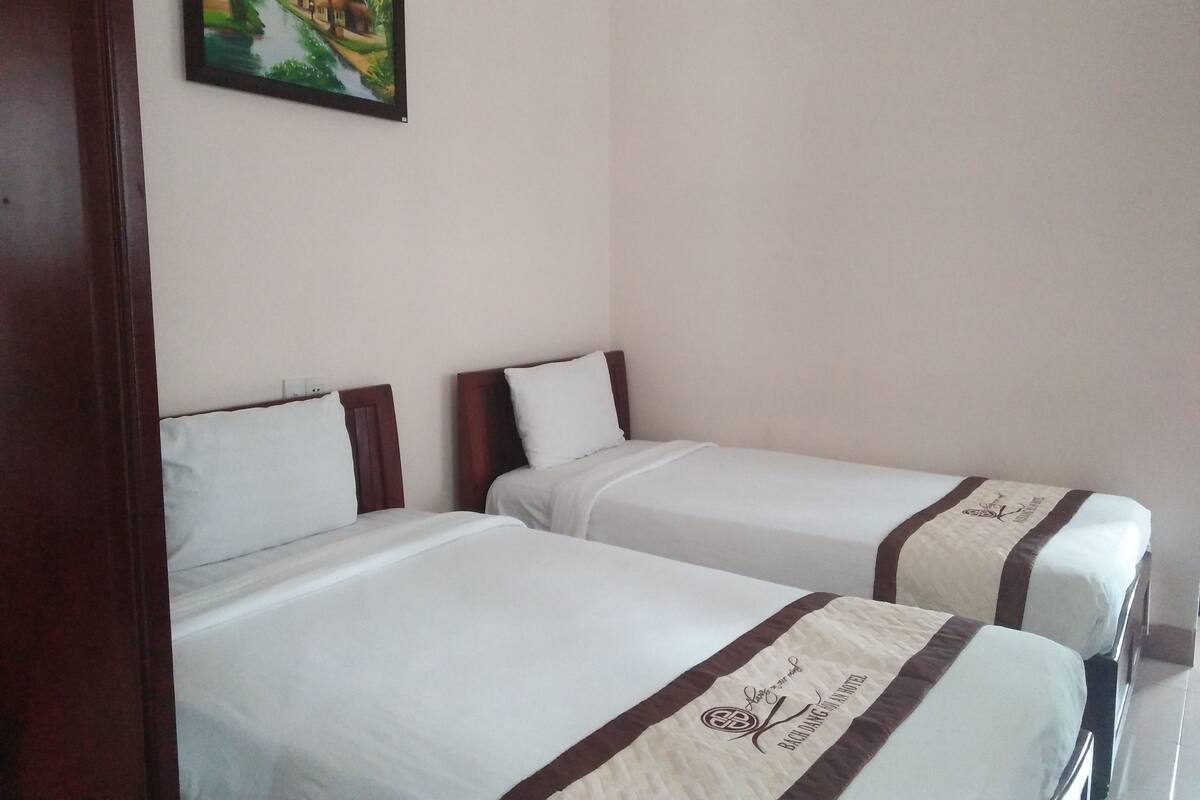 Two single beds with white linens and brown headboards, a painting of a tropical scene on the wall, and a wooden door frame.