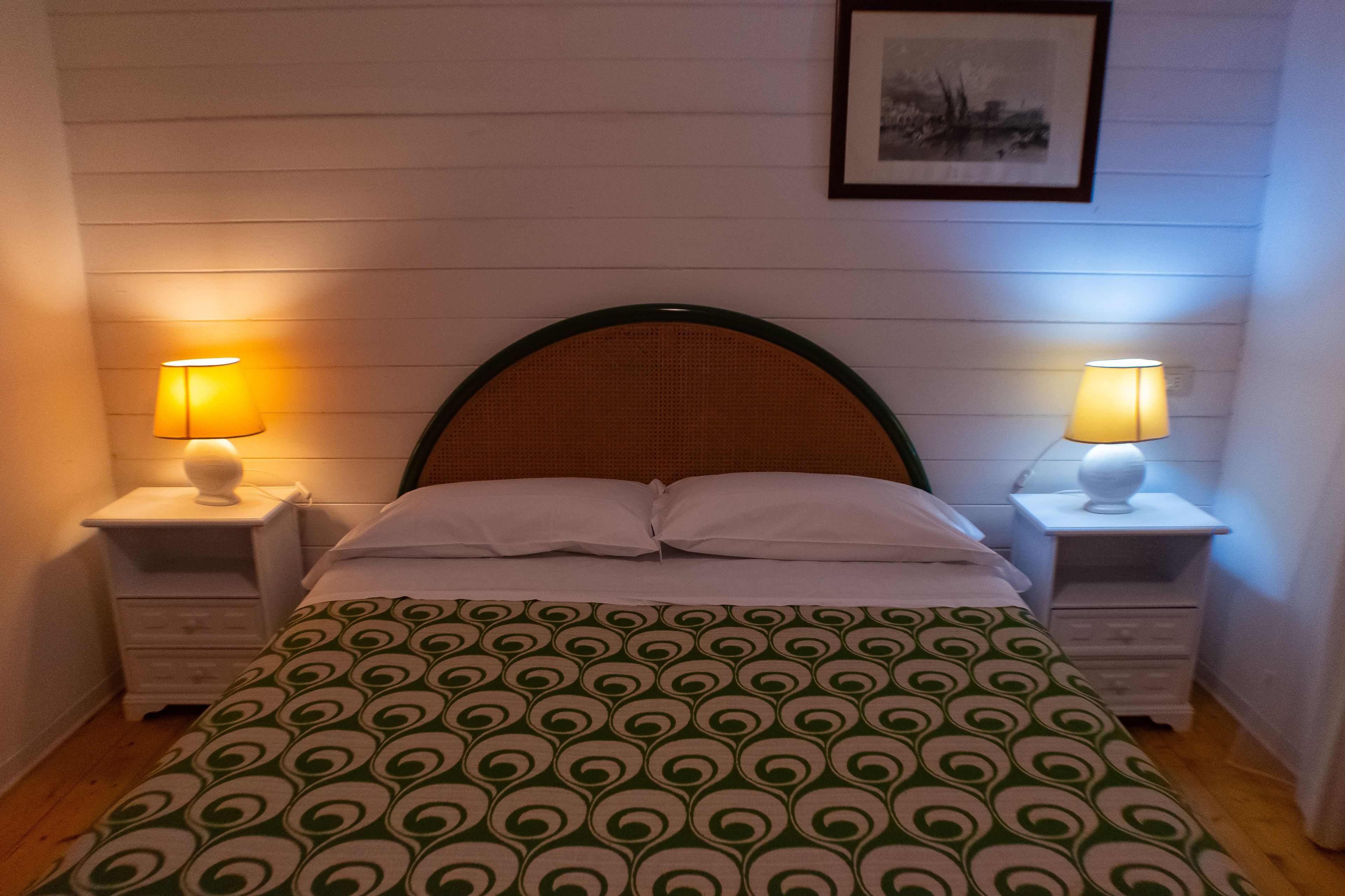Un dormitorio con una cama, dos mesitas de noche con lámparas, un cuadro enmarcado en la pared y una colcha estampada.
