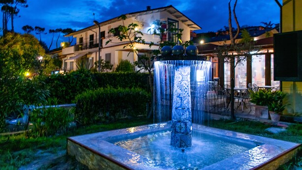 Une fontaine dont l’eau jaillit en cascade le long d’une colonne, entourée de verdure, avec un bâtiment en arrière-plan.