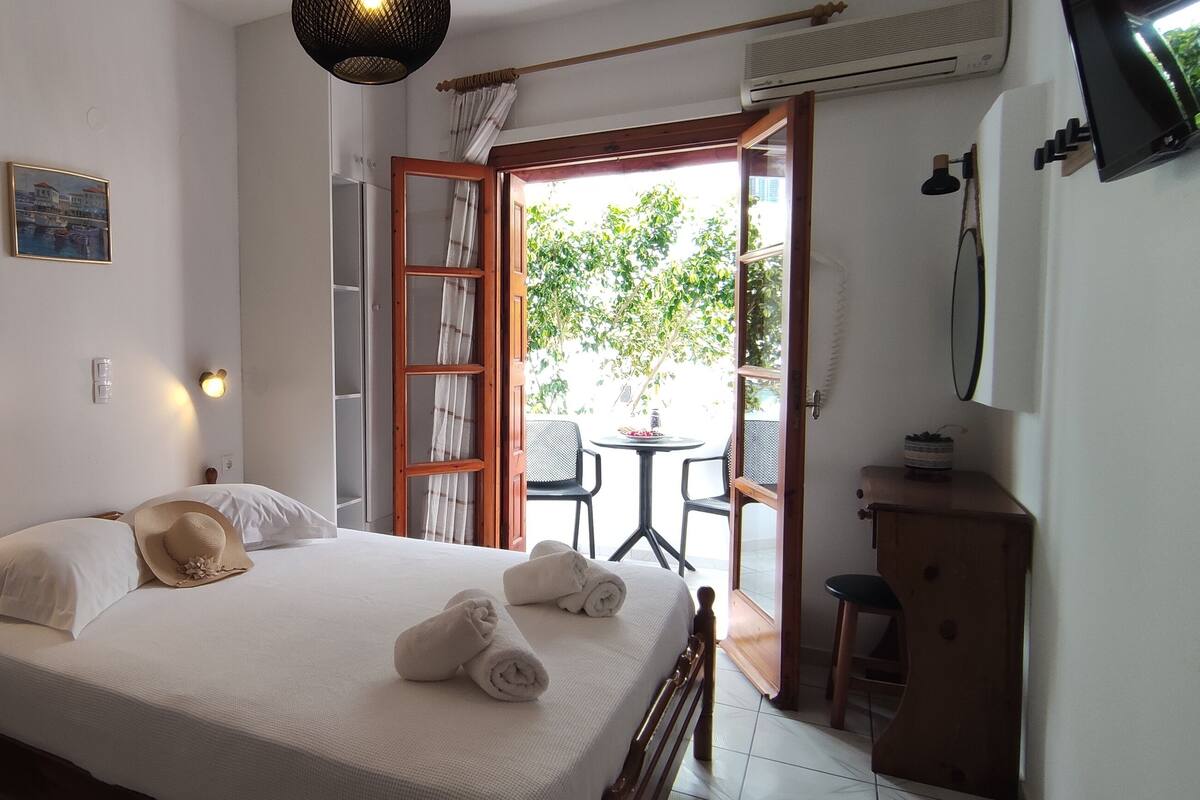 A neatly made bed with white linens and towels, a bedside table with a book, a framed picture on the wall, and a view of the outdoors through an open door.