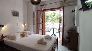 A neatly made bed with white linens and towels, a bedside table with a book, a framed picture on the wall, and a view of the outdoors through an open door.