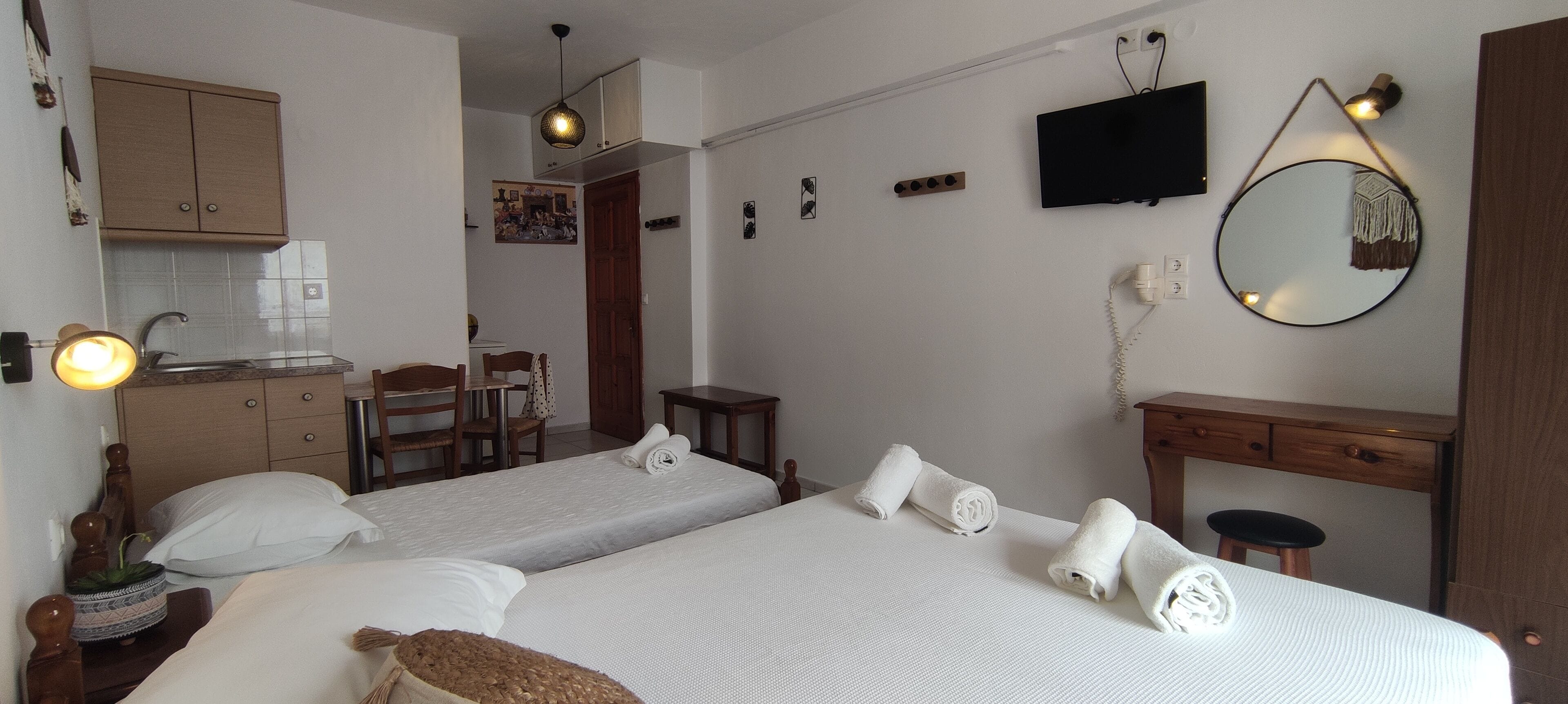 Habitación de hotel con dos camas, un escritorio, una silla, una kitchenette pequeña y un televisor montado en la pared.