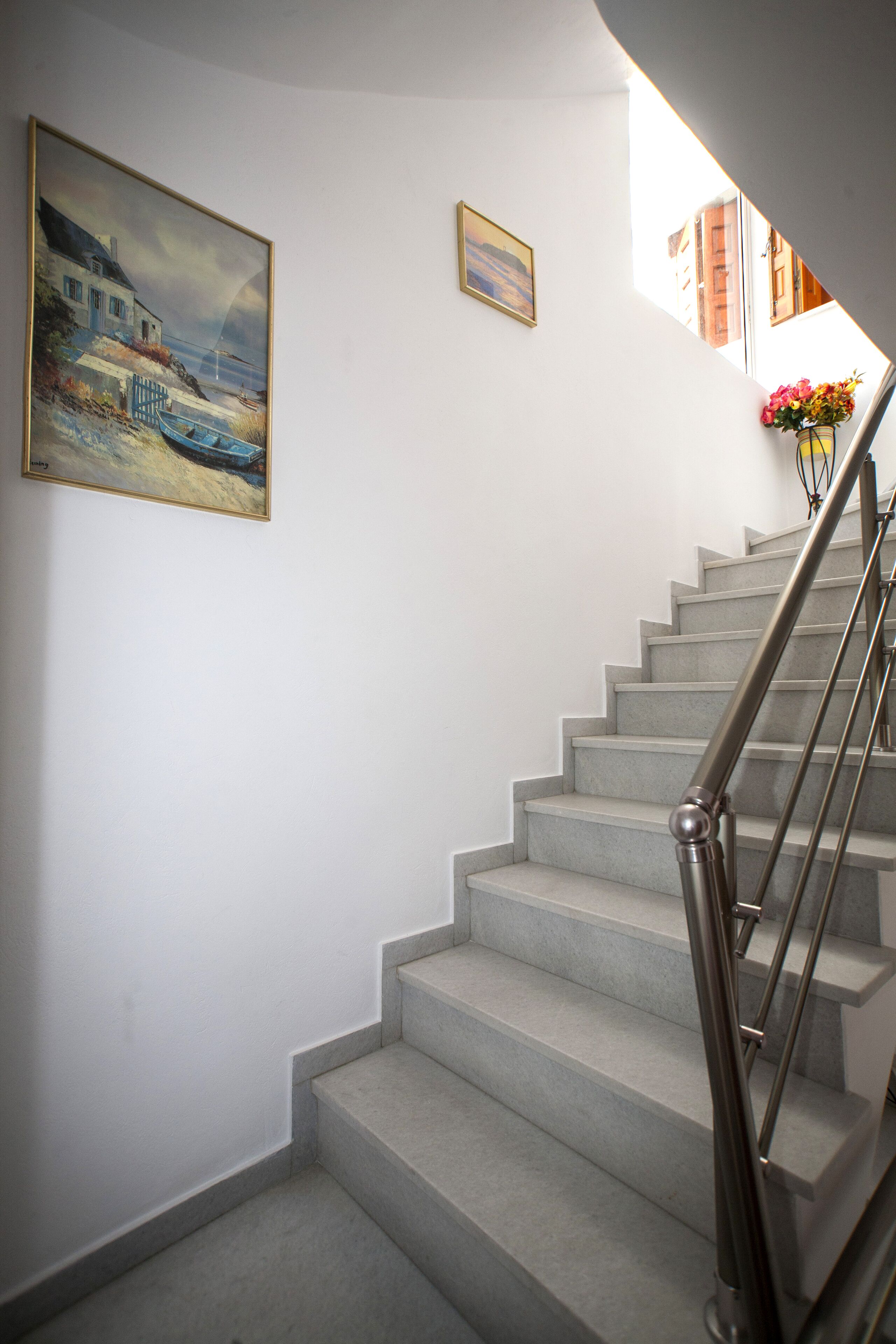 Una escalera con barandilla metálica, un cuadro de una escena costera y un florero con flores en la pared.