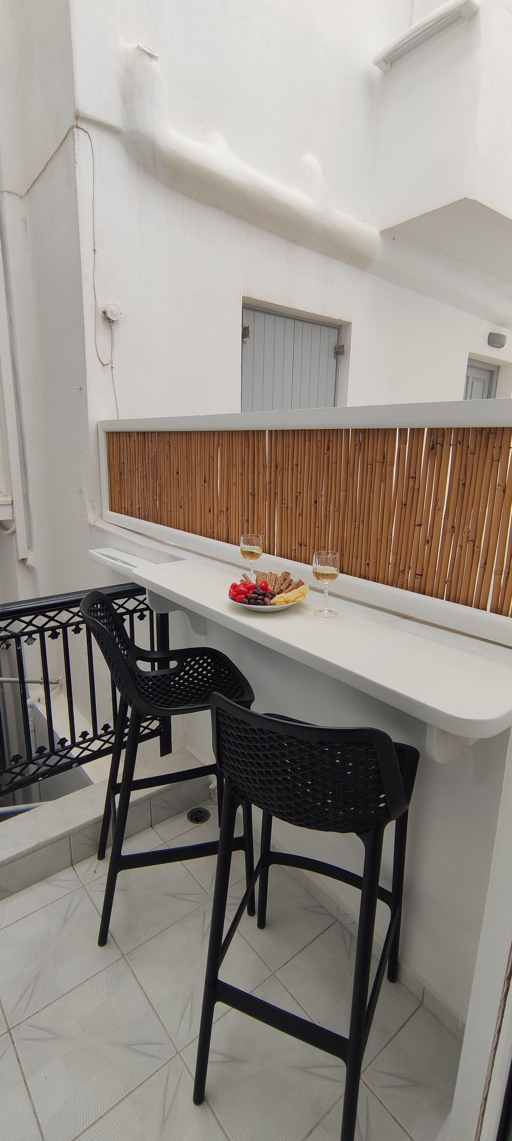 Uma pequena área de refeições ao ar livre, com uma mesa branca e duas cadeiras pretas, um painel de madeira e uma pequena janela.