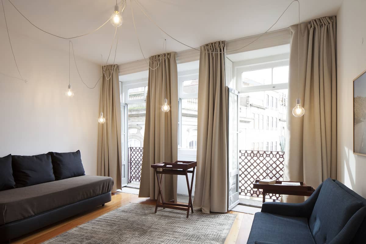 A modern living room with a sofa, a small table, and a chair. There are curtains on the windows, and a balcony is visible.