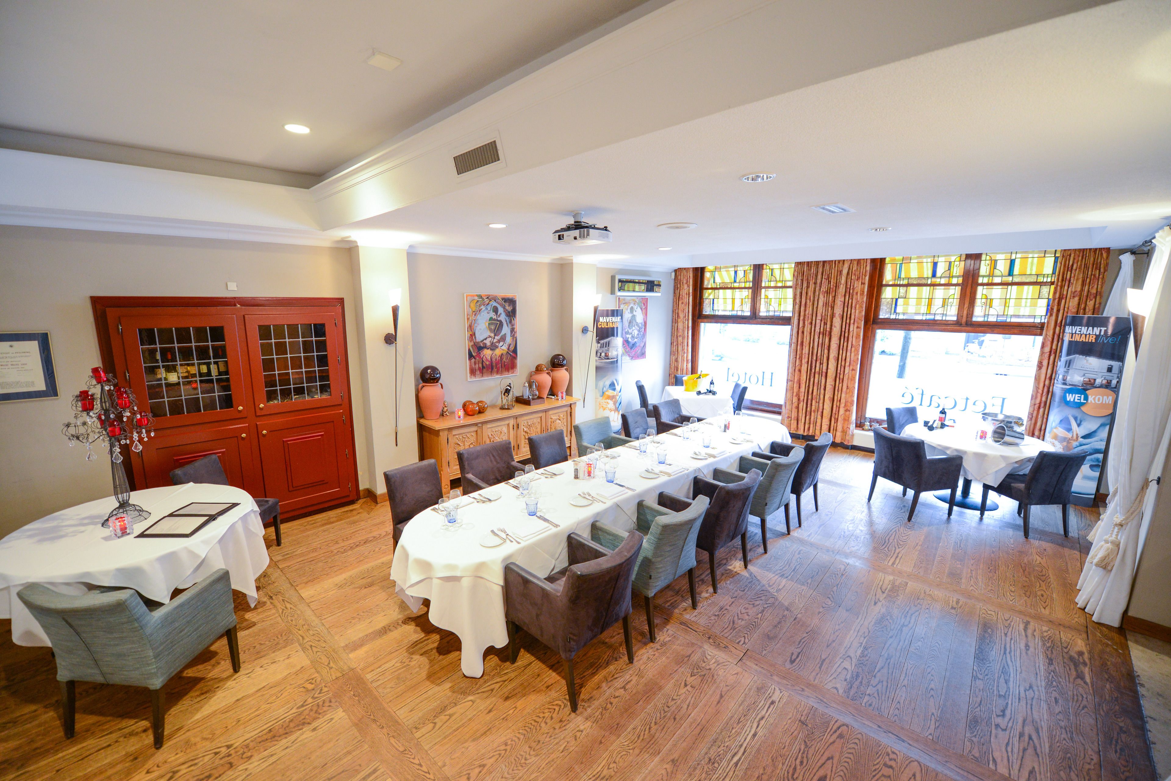 Uma sala de jantar ampla com uma mesa longa posta para várias pessoas, piso de madeira e vista através de grandes janelas.