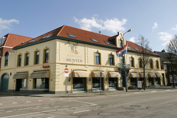 Un bâtiment sur lequel on peut lire l’inscription « MUNTEN » et qui possède plusieurs devantures de magasins avec des auvents.