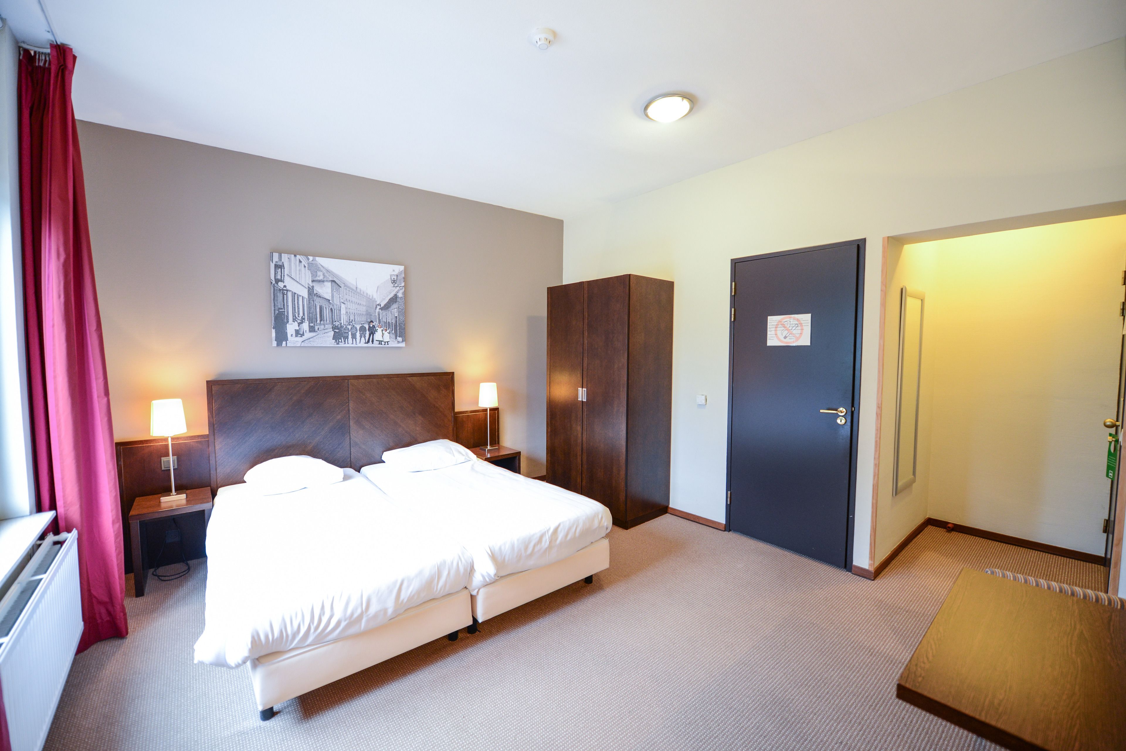 A hotel room with a bed, two bedside tables with lamps, a wardrobe, a door with a 'No Entry' sign, and a wall-mounted picture.