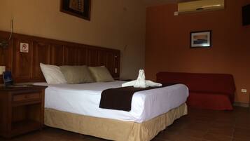 Habitación de hotel con cabecera de madera, cama con ropa blanca, sofá rojo y aire acondicionado instalado en la pared.