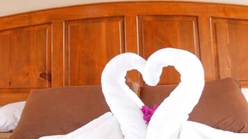 A bed with a wooden headboard, a framed artwork above it, and a towel arranged in a heart shape on the bed.