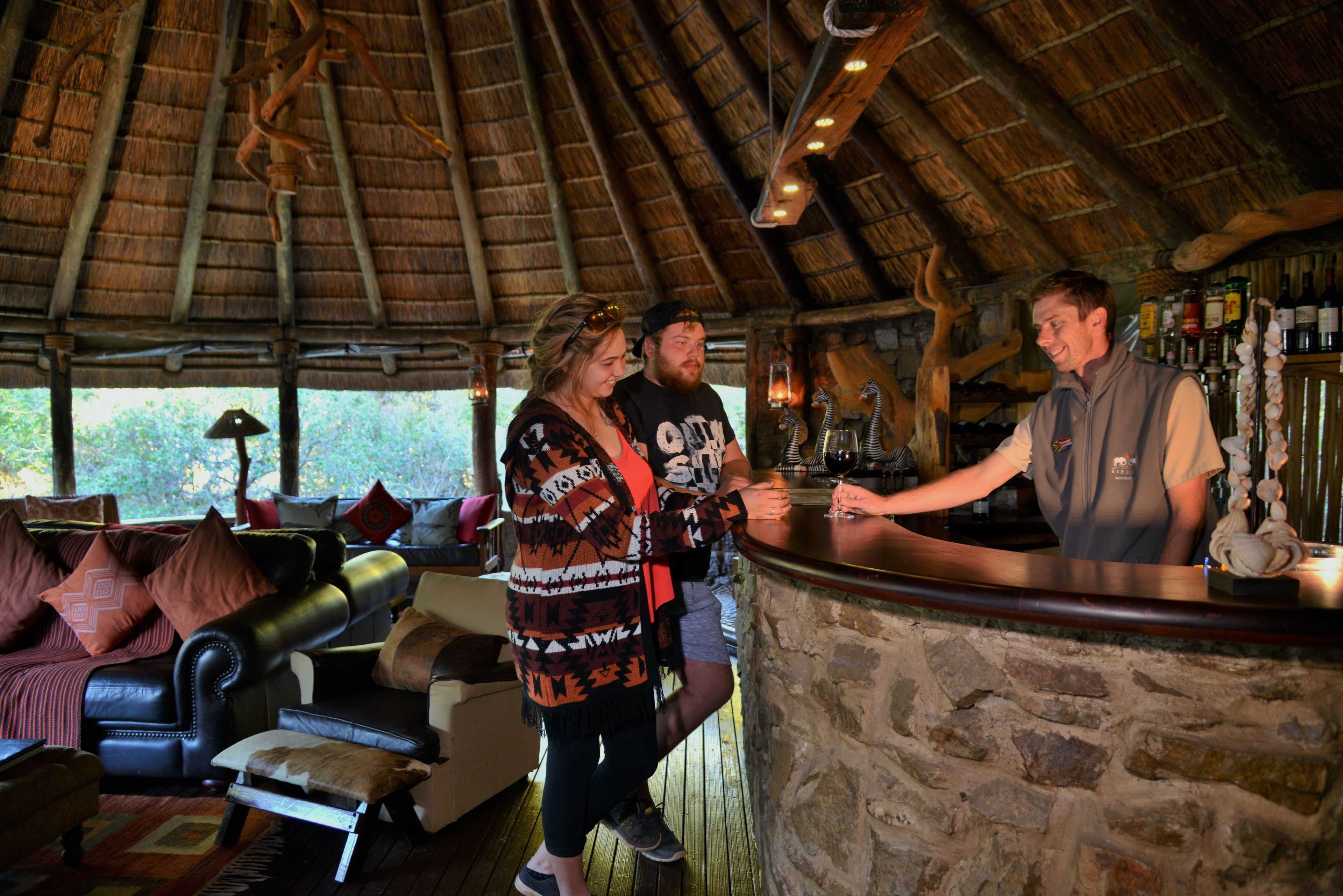 A rustic bar area with a stone counter, wooden furniture, and a thatched roof.