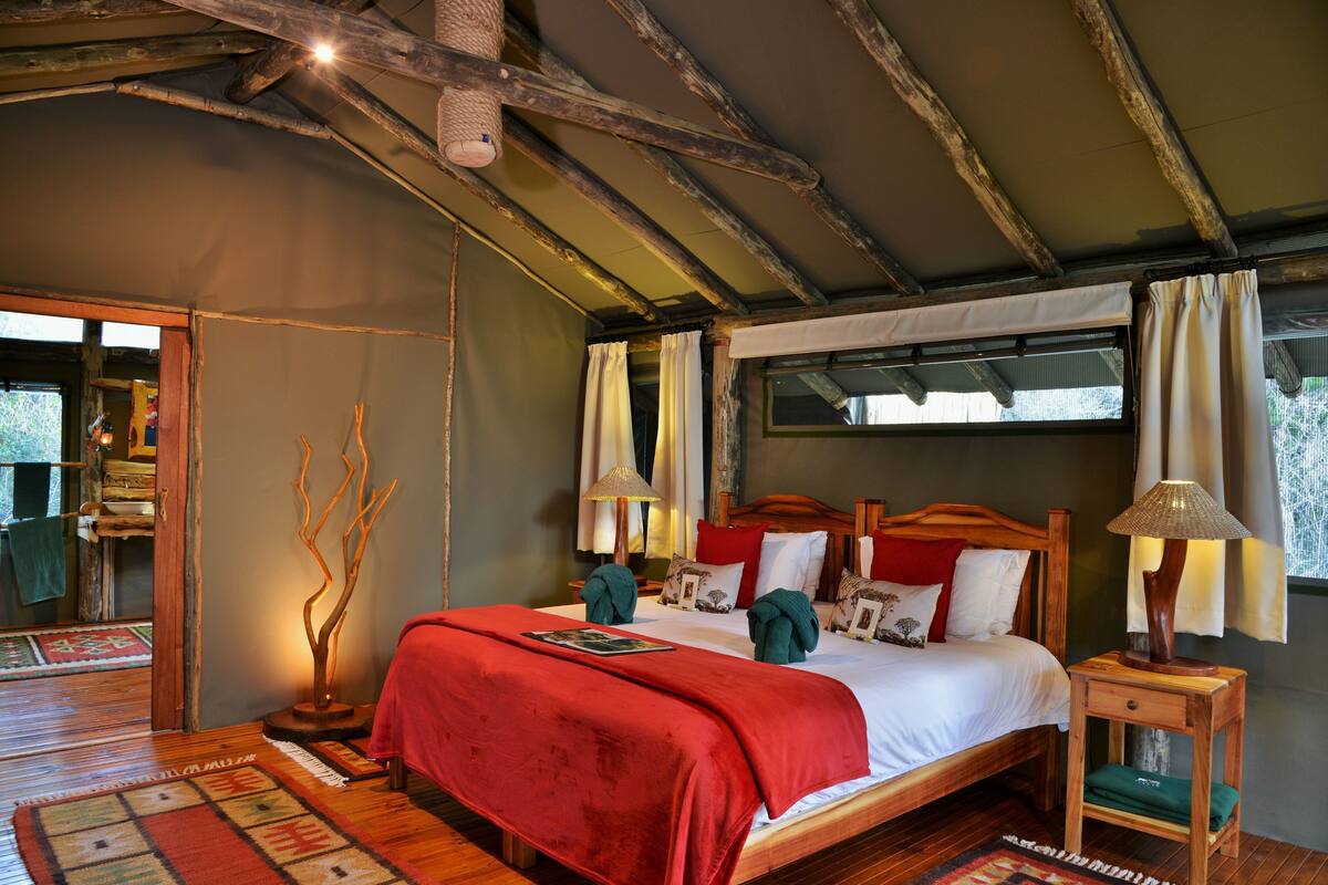 A bedroom with a wooden bed, red bedding, a nightstand with a lamp, and a view of the outdoors.