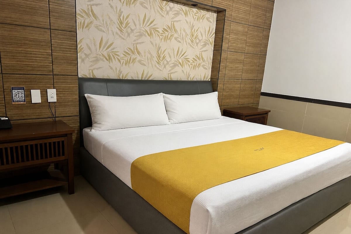 Een hotelkamer met een bed, houten nachtkastjes, een hoofdbord met een bladmotief en een geel sierkussen.