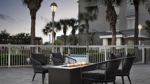 An outdoor seating area with a fire feature, surrounded by a white fence, palm trees, and a building in the background. - Homewood Suites by Hilton Orlando Airport (Orlando)