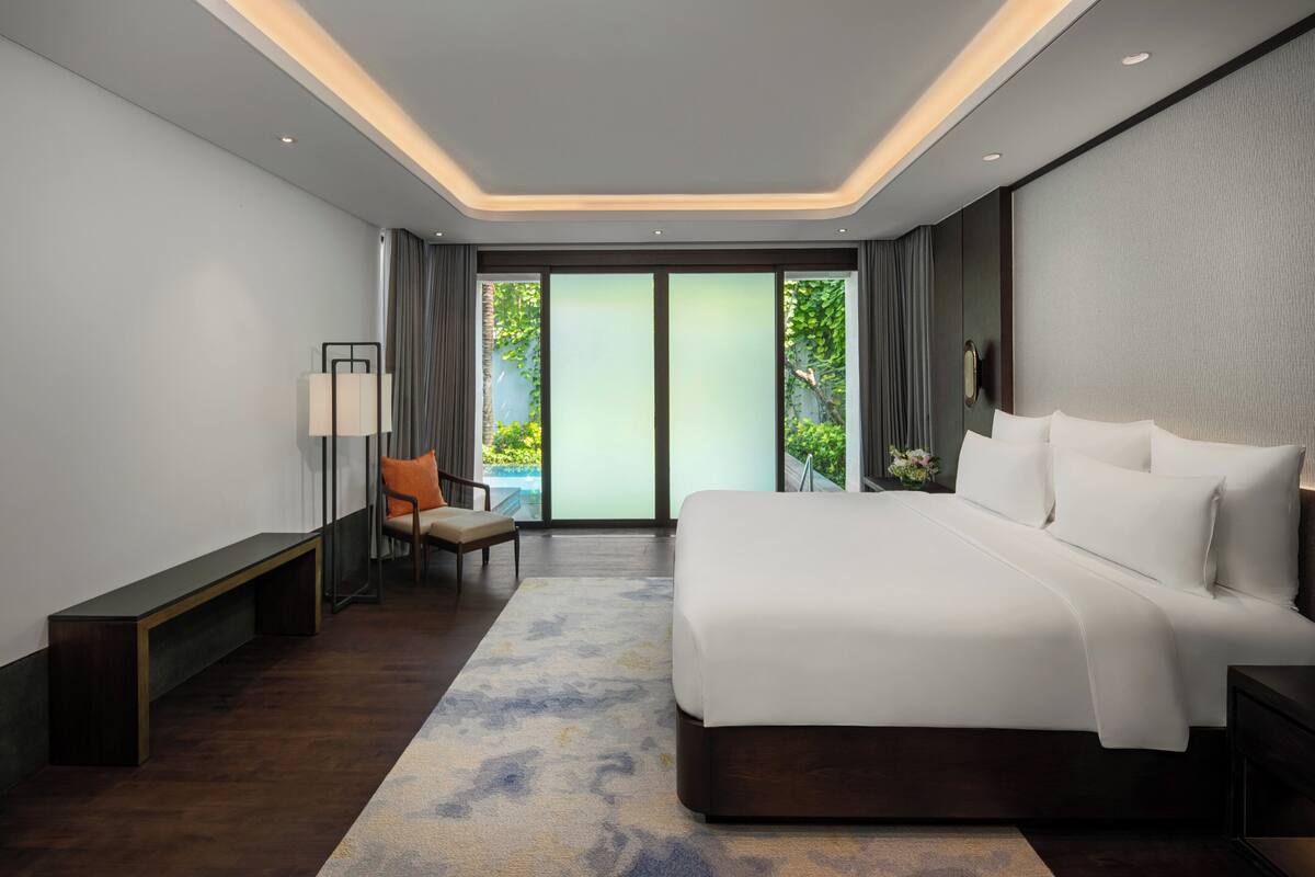 Une chambre d’hôtel moderne avec un grand lit, un banc, une chaise et une vue sur la verdure à travers les portes vitrées.