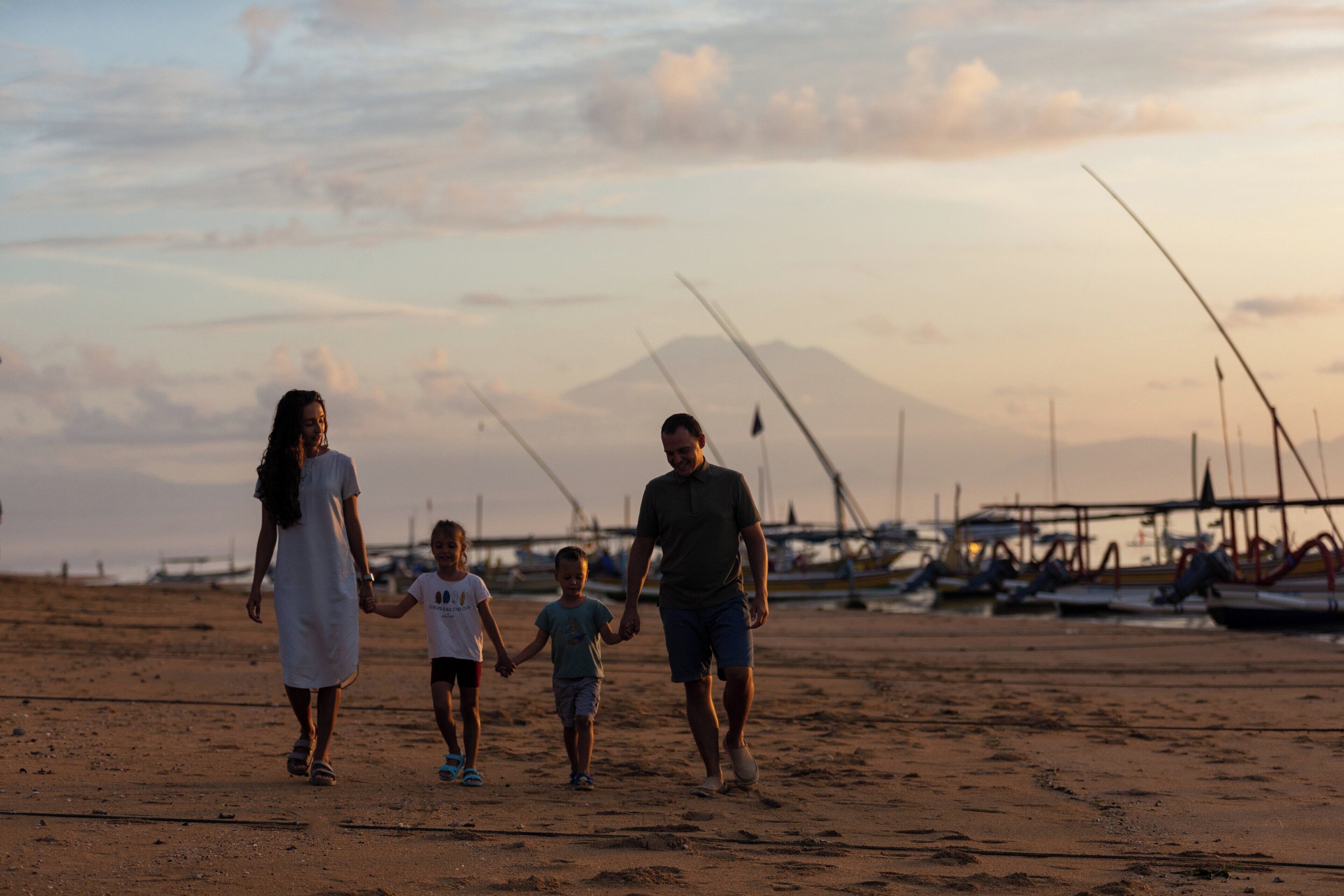 Una familia caminando sobre una playa arenosa con barcos al fondo.